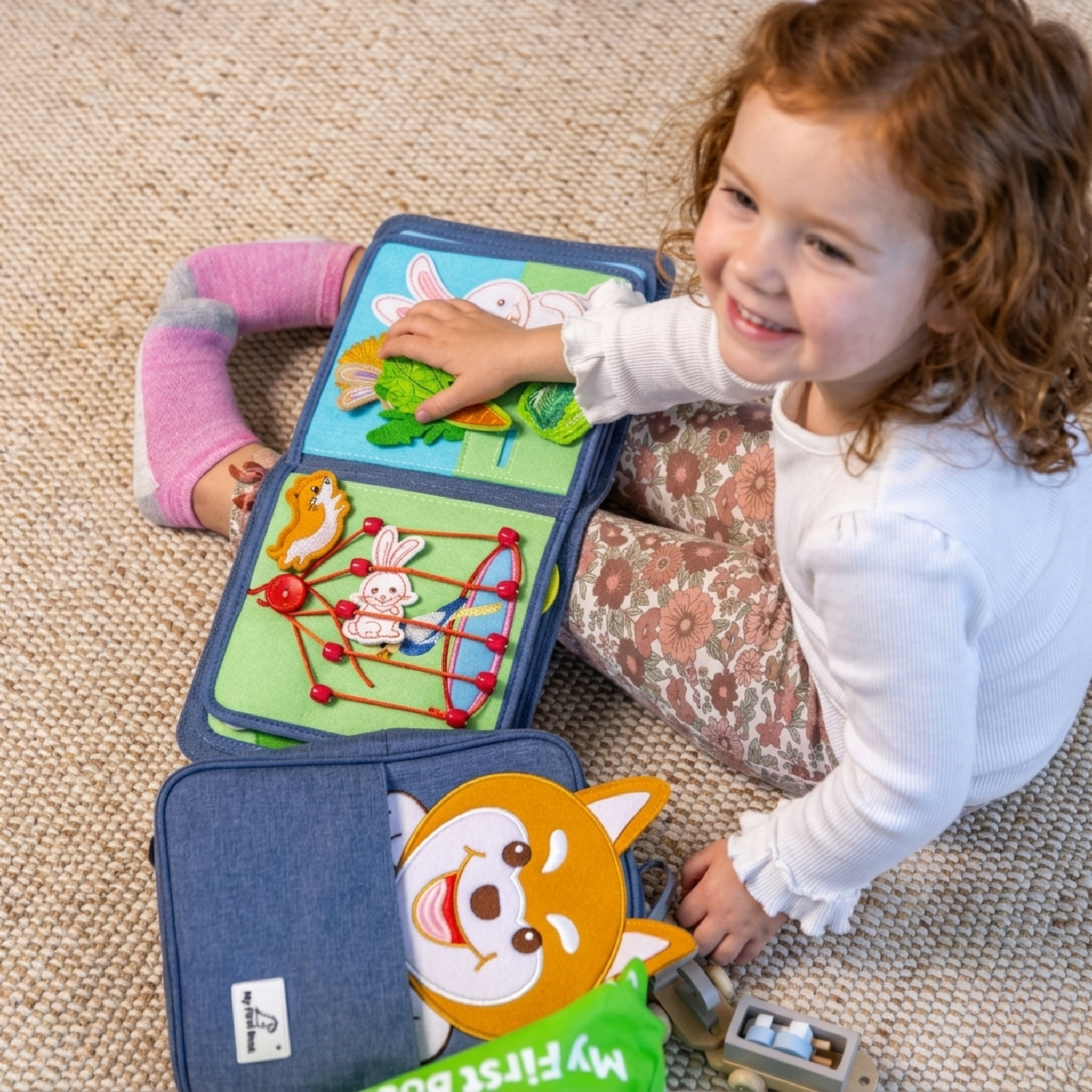 Ein Kind sitzt lächelnd auf einem Teppich mit dem Montessori Quiet Book Pet Friends von My First Book auf dem Schoß und den passenden Rucksack neben ihr liegend.