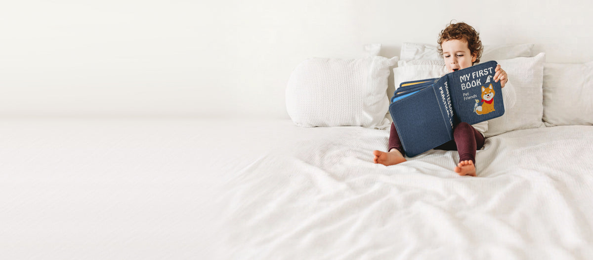Kleinkind sitzt auf einem weißen Bett und liest im My First Book Pet Friends Quiet Book