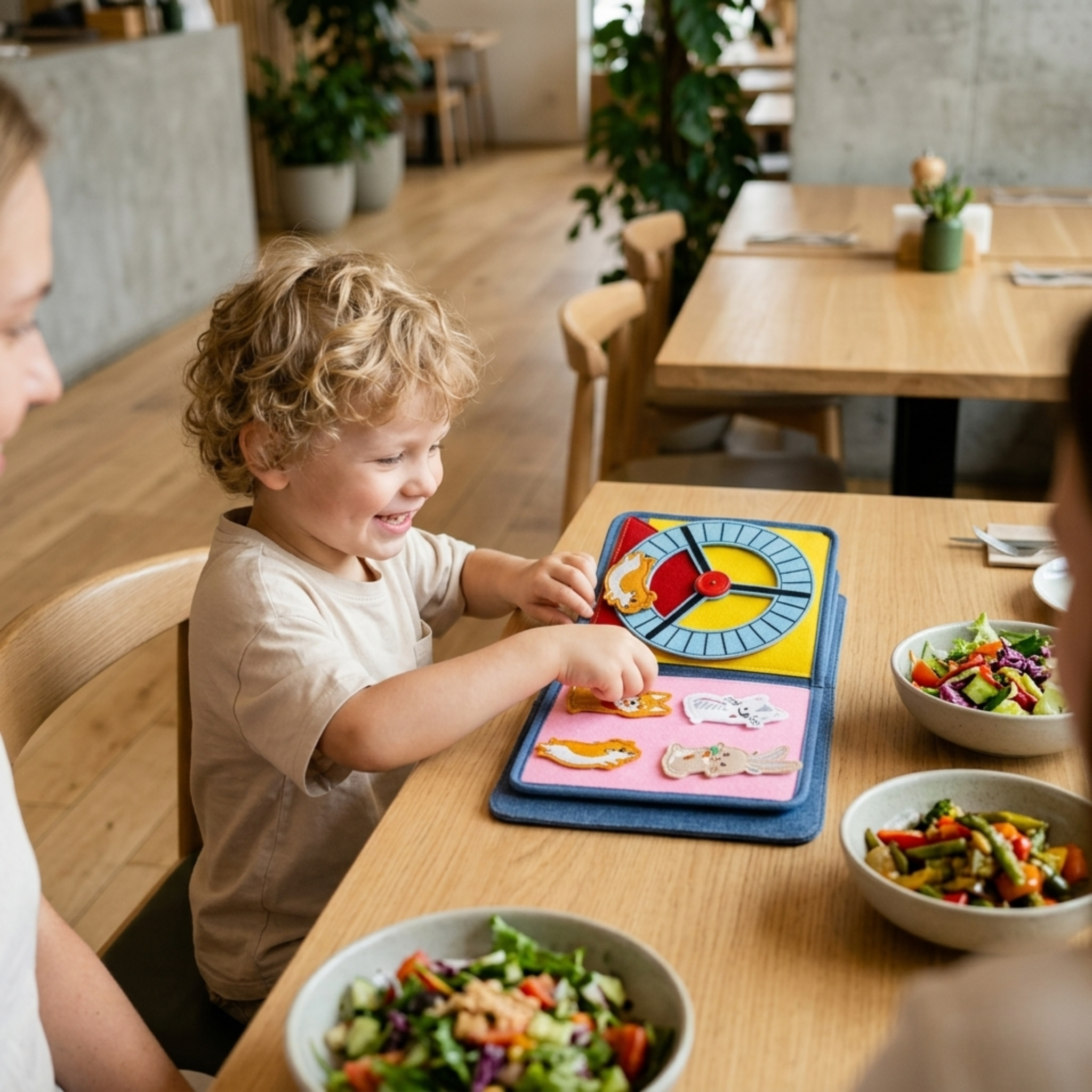 Ein lächelndes Kind spielt im Restaurant mit dem 'My First Book' Montessori Quiet Book aus Stoff. Zu sehen ist die Seite mit einem bunten Filz-Drehrad und abnehmbaren Tierfiguren wie Hund und Hase. Auf dem Tisch im Hintergrund ist gesundes Essen in Schüsseln angerichtet.